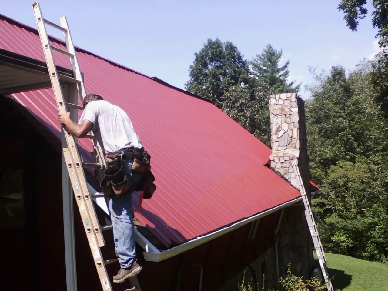 Roofer on a ladder beside a red metal roof and stone chimney — Soffit & Fascia Repair by Roofing Brothers in Katy